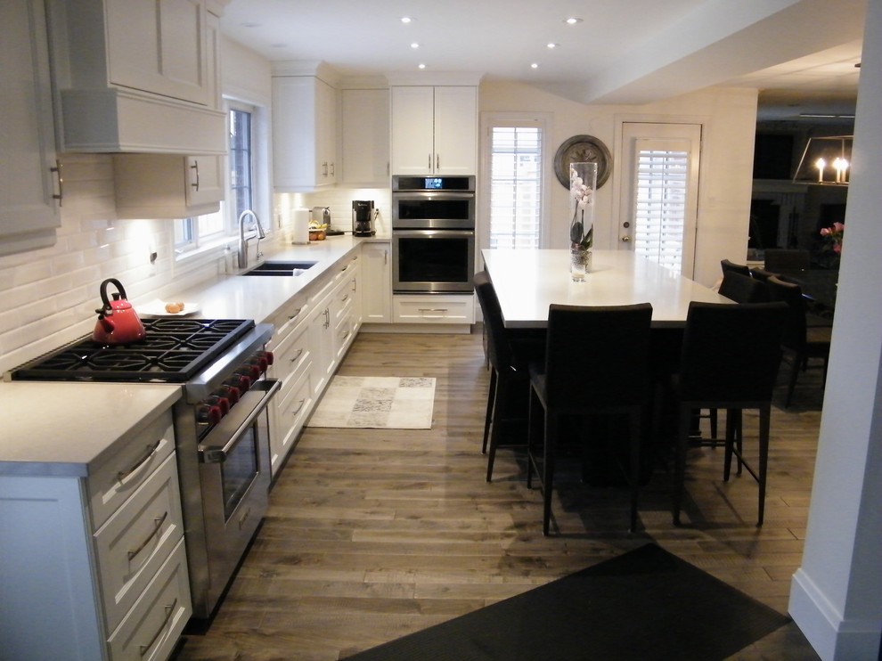 Idéer för att renovera ett mellanstort funkis vit vitt kök, med en dubbel diskho, luckor med infälld panel, vita skåp, vitt stänkskydd, stänkskydd i tunnelbanekakel, rostfria vitvaror, mellanmörkt trägolv, en köksö, bänkskiva i kvarts och brunt golv