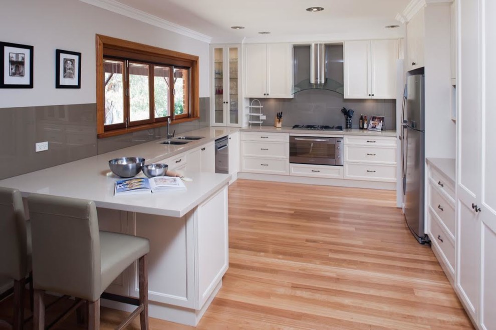 Inspiration for a medium sized classic u-shaped enclosed kitchen in Los Angeles with a double-bowl sink, recessed-panel cabinets, white cabinets, engineered stone countertops, grey splashback, glass tiled splashback, stainless steel appliances, light hardwood flooring, a breakfast bar and beige floors.