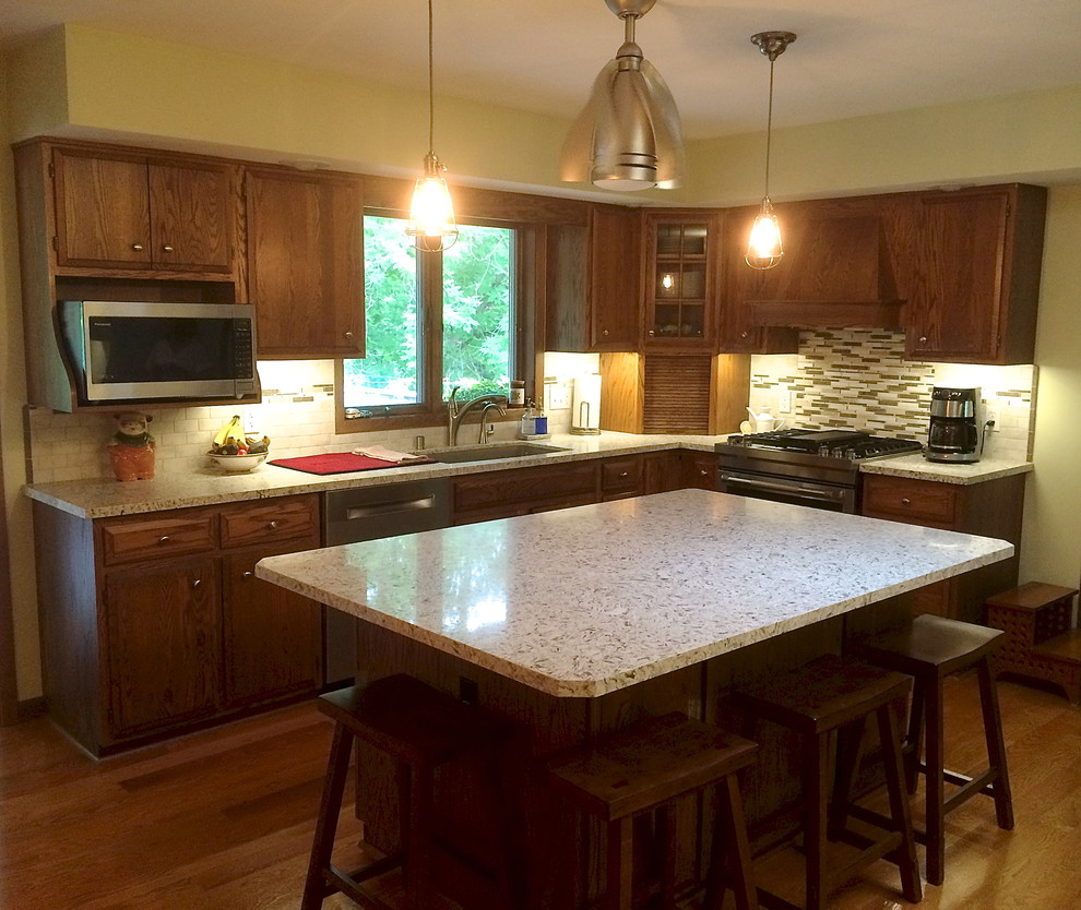 This is an example of a medium sized eclectic l-shaped kitchen/diner in Milwaukee with a submerged sink, raised-panel cabinets, medium wood cabinets, engineered stone countertops, multi-coloured splashback, glass tiled splashback, stainless steel appliances, medium hardwood flooring and an island.