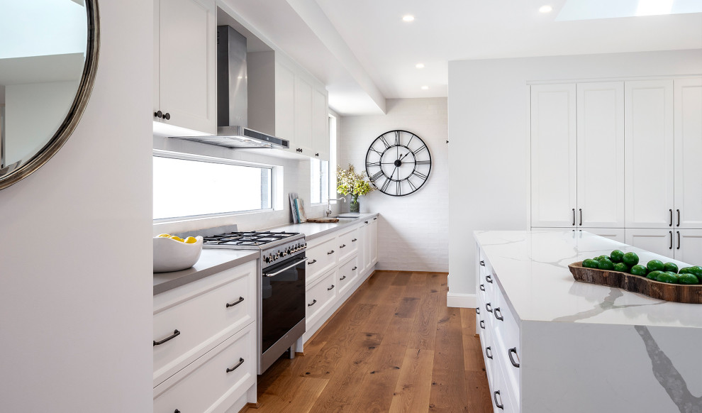 Foto på ett stort maritimt grå l-kök, med en undermonterad diskho, luckor med infälld panel, vita skåp, vitt stänkskydd, integrerade vitvaror, mellanmörkt trägolv, en köksö och brunt golv