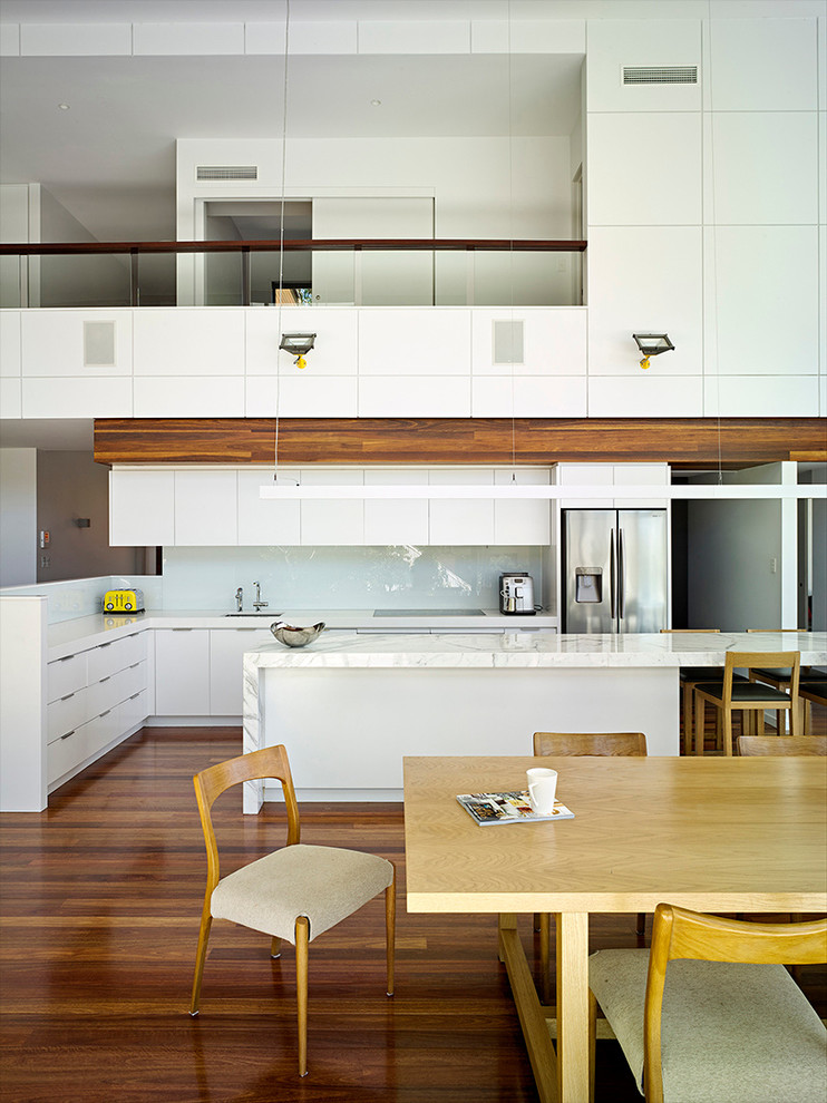 Zweizeilige, Mittelgroße Moderne Wohnküche mit Unterbauwaschbecken, flächenbündigen Schrankfronten, weißen Schränken, Marmor-Arbeitsplatte, Küchenrückwand in Weiß, Glasrückwand, braunem Holzboden, Kücheninsel und Küchengeräten aus Edelstahl in Brisbane