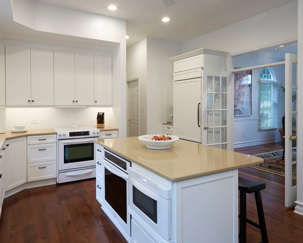 Example of a mid-sized transitional l-shaped dark wood floor eat-in kitchen design in Baltimore with an undermount sink, shaker cabinets, white cabinets, solid surface countertops, white backsplash, white appliances and an island