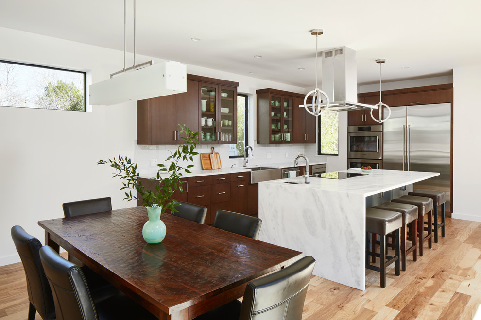 Contemporary l-shaped kitchen/diner in Austin with a belfast sink, flat-panel cabinets, dark wood cabinets, white splashback, stainless steel appliances, light hardwood flooring, an island, beige floors and white worktops.