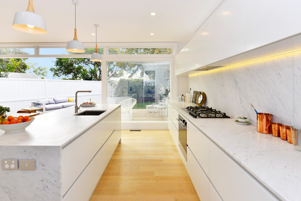 This is an example of a medium sized contemporary galley kitchen/diner in Auckland with a submerged sink, white cabinets, marble worktops, white splashback, stainless steel appliances, light hardwood flooring, an island and flat-panel cabinets.