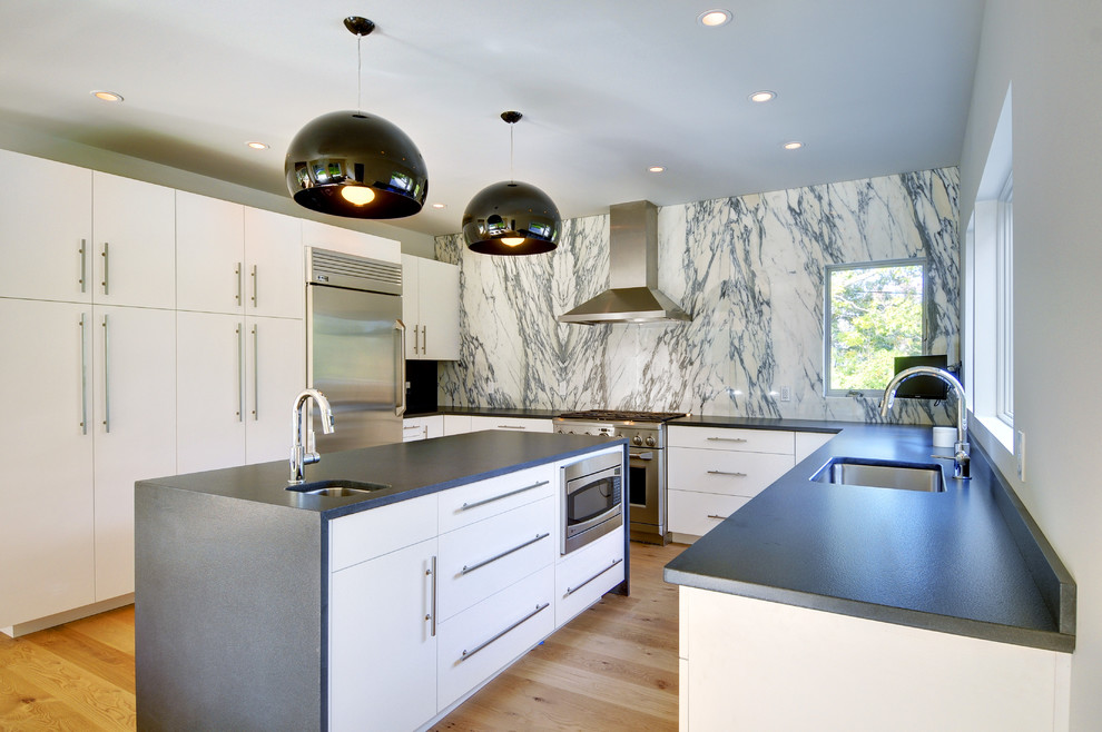 Modern inredning av ett stort kök, med en undermonterad diskho, släta luckor, vita skåp, granitbänkskiva, stänkskydd i sten, rostfria vitvaror, vitt stänkskydd, ljust trägolv och en köksö