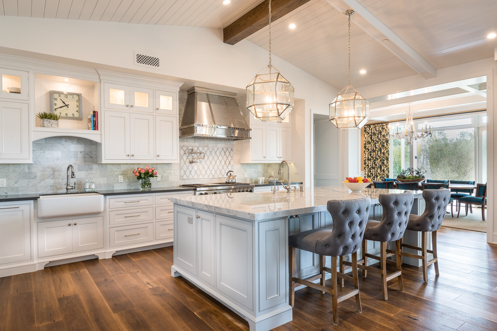 Inspiration for a coastal kitchen in Los Angeles with a belfast sink, recessed-panel cabinets, white cabinets, grey splashback, stainless steel appliances, medium hardwood flooring and an island.
