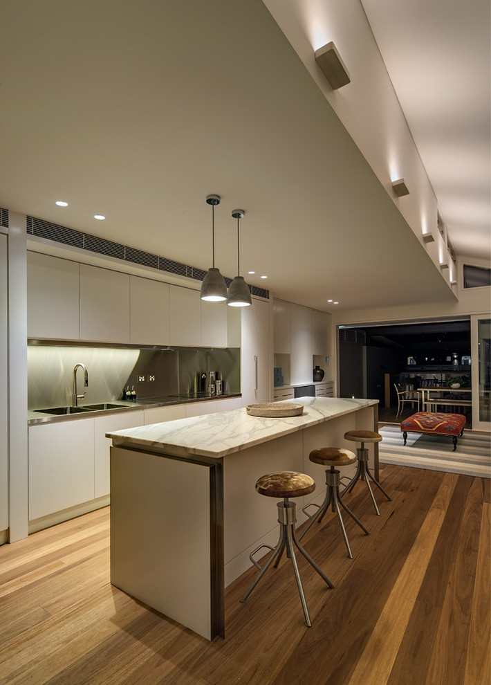 Modern inredning av ett mellanstort kök, med en nedsänkt diskho, släta luckor, vita skåp, bänkskiva i rostfritt stål, stänkskydd med metallisk yta, rostfria vitvaror, mellanmörkt trägolv och en köksö