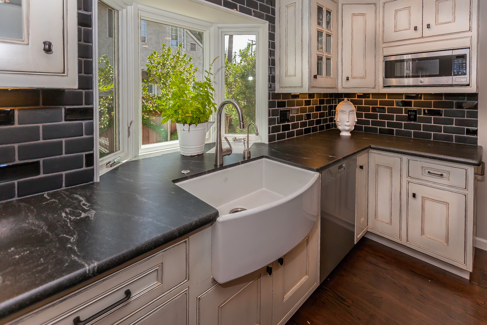 Mid-sized cottage u-shaped dark wood floor and brown floor eat-in kitchen photo in San Francisco with a farmhouse sink, recessed-panel cabinets, white cabinets, marble countertops, black backsplash, subway tile backsplash, stainless steel appliances and an island