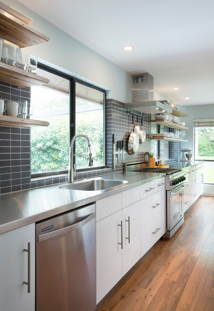 Bild på ett funkis kök, med en integrerad diskho, släta luckor, vita skåp, bänkskiva i rostfritt stål, grått stänkskydd, rostfria vitvaror och mellanmörkt trägolv