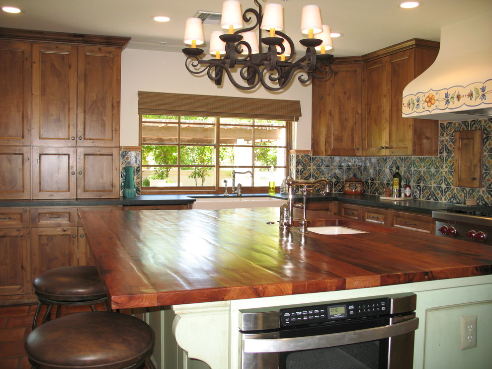 Large southwest l-shaped painted wood floor eat-in kitchen photo in Phoenix with a farmhouse sink, recessed-panel cabinets, distressed cabinets, wood countertops, multicolored backsplash, terra-cotta backsplash, paneled appliances and an island