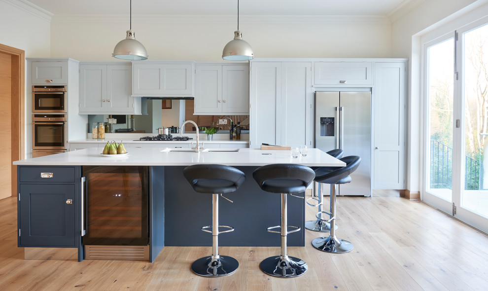Imagen de cocina clásica renovada con armarios estilo shaker, puertas de armario blancas, electrodomésticos de acero inoxidable, suelo de madera clara y una isla