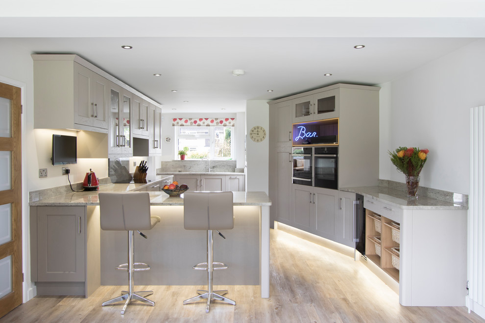 Medium sized traditional u-shaped enclosed kitchen in Other with a submerged sink, recessed-panel cabinets, grey cabinets, granite worktops, grey splashback, a breakfast bar, black appliances, light hardwood flooring and beige floors.