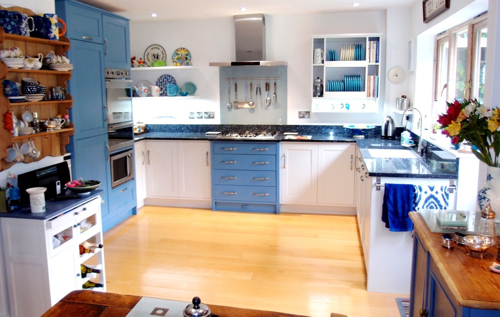 Design ideas for a large contemporary u-shaped open plan kitchen in Cornwall with a double-bowl sink, shaker cabinets, blue cabinets, granite worktops, metallic splashback, glass sheet splashback, stainless steel appliances, light hardwood flooring and an island.