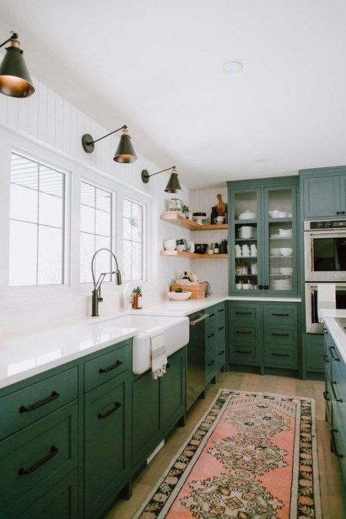 Stylish best kitchen cabinet colors Canada with deep green shaker cabinets, white farmhouse sink, glass-front upper cabinets, and brass wall sconces, highlighting one of the best kitchen cabinet color choices in Canada.