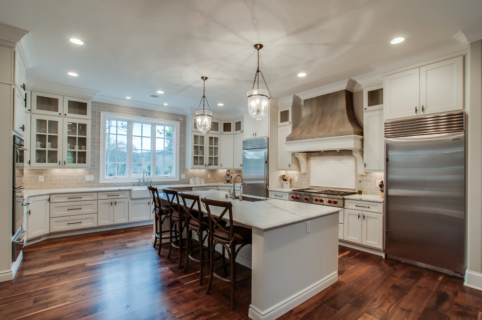 Large cottage chic u-shaped medium tone wood floor eat-in kitchen photo in Wichita with a farmhouse sink, recessed-panel cabinets, white cabinets, marble countertops, beige backsplash, subway tile backsplash, stainless steel appliances and an island