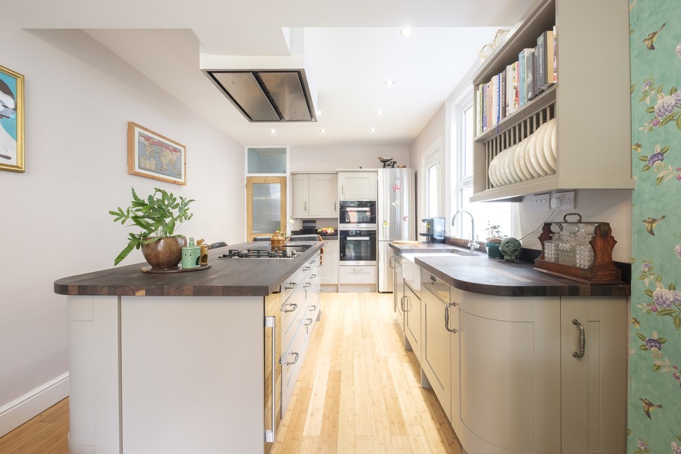 This is an example of a medium sized traditional galley open plan kitchen in Kent with a belfast sink, shaker cabinets, beige cabinets, wood worktops, brown splashback, wood splashback, stainless steel appliances, medium hardwood flooring, an island and multi-coloured floors.