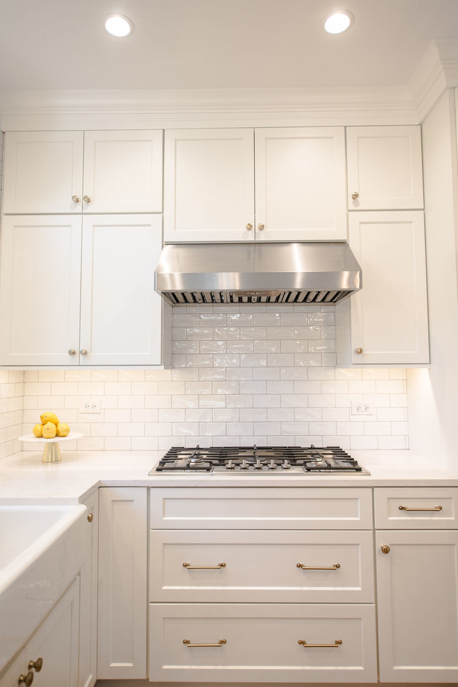 This is an example of a medium sized traditional u-shaped kitchen pantry in Chicago with a belfast sink, flat-panel cabinets, white cabinets, engineered stone countertops, ceramic splashback, stainless steel appliances, medium hardwood flooring and an island.