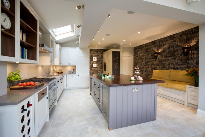 Example of a large ornate l-shaped travertine floor eat-in kitchen design in London with an undermount sink, recessed-panel cabinets, white cabinets, wood countertops, beige backsplash, glass sheet backsplash, stainless steel appliances and an island