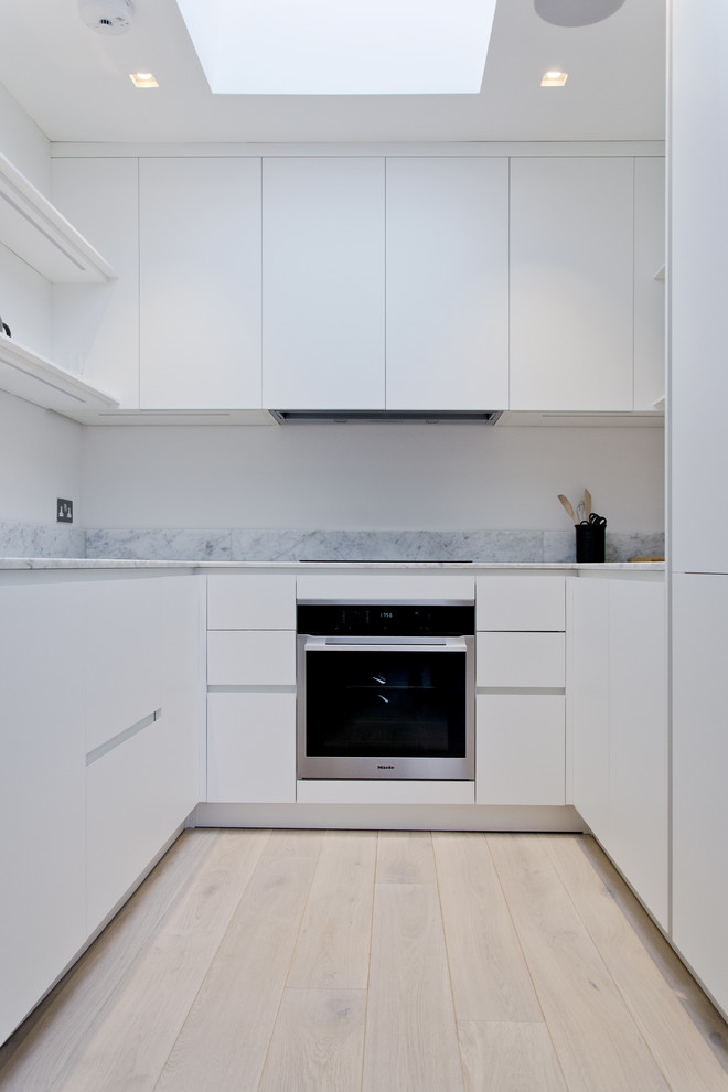 Modern inredning av ett avskilt, mellanstort u-kök, med en nedsänkt diskho, släta luckor, vita skåp, marmorbänkskiva, grått stänkskydd, rostfria vitvaror och ljust trägolv