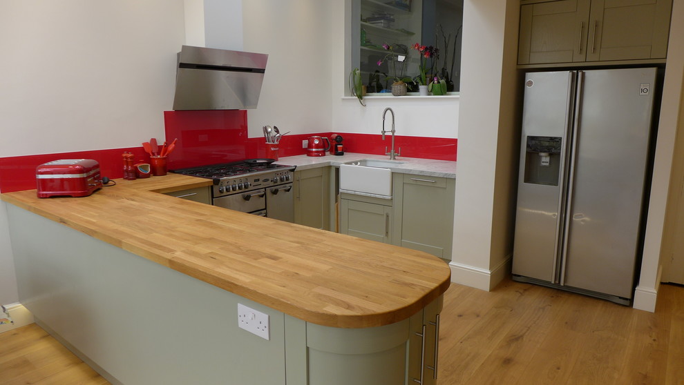 Inspiration for a medium sized contemporary u-shaped open plan kitchen in Other with a belfast sink, shaker cabinets, green cabinets, wood worktops, red splashback, glass sheet splashback, stainless steel appliances and light hardwood flooring.
