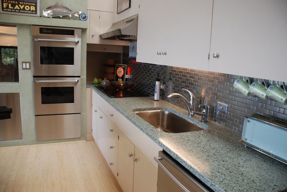 Inredning av ett modernt kök och matrum, med en undermonterad diskho, vita skåp, bänkskiva i återvunnet glas, grått stänkskydd, stänkskydd i metallkakel, rostfria vitvaror och ljust trägolv