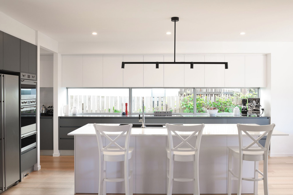 Design ideas for a contemporary l-shaped kitchen in Gold Coast - Tweed with a built-in sink, flat-panel cabinets, grey cabinets, window splashback, stainless steel appliances, light hardwood flooring, an island, beige floors and grey worktops.
