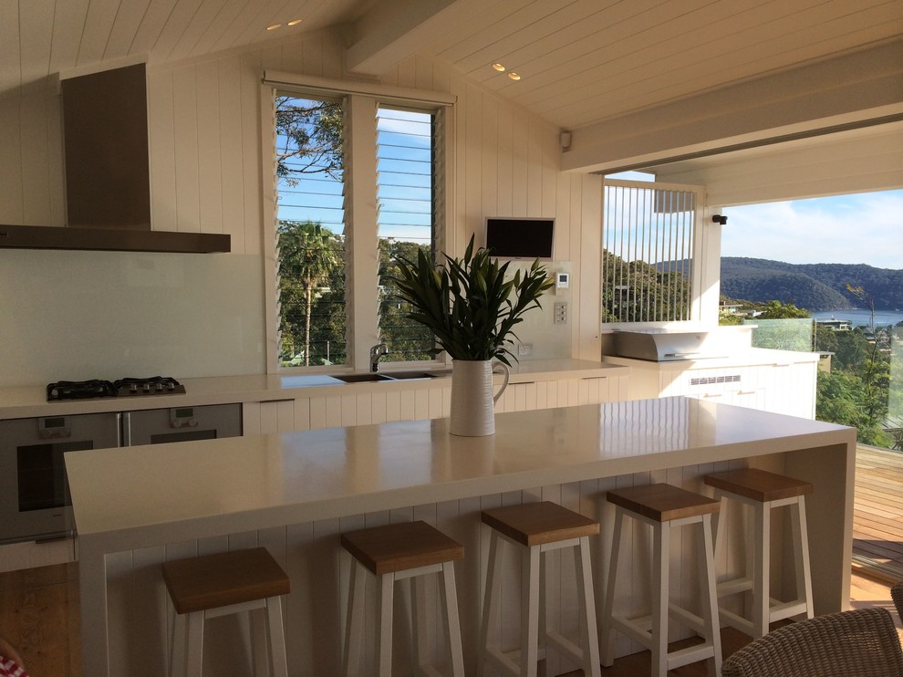 Inspiration for a large coastal galley open plan kitchen in Sydney with a double-bowl sink, white cabinets, composite countertops, white splashback, glass sheet splashback, stainless steel appliances, light hardwood flooring and an island.