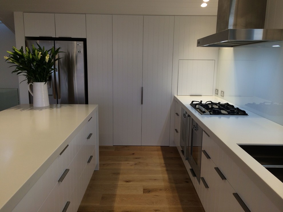 Inspiration for a large coastal galley light wood floor open concept kitchen remodel in Sydney with a double-bowl sink, white cabinets, solid surface countertops, white backsplash, glass sheet backsplash, stainless steel appliances and an island