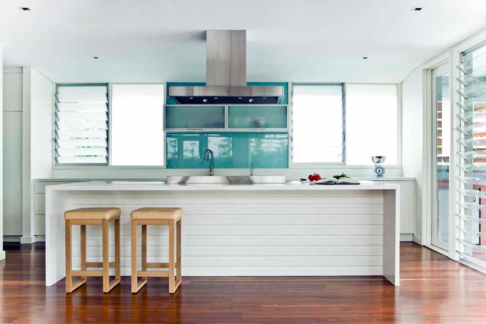 Example of a beach style galley medium tone wood floor kitchen design in Sydney with white cabinets, blue backsplash, glass sheet backsplash and an island