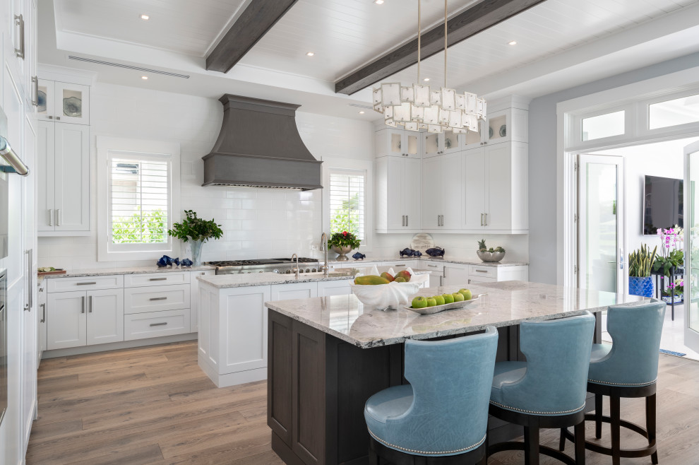 This is an example of a coastal kitchen in Other with a belfast sink, recessed-panel cabinets, white cabinets, white splashback, metro tiled splashback, stainless steel appliances, medium hardwood flooring, multiple islands, brown floors and exposed beams.