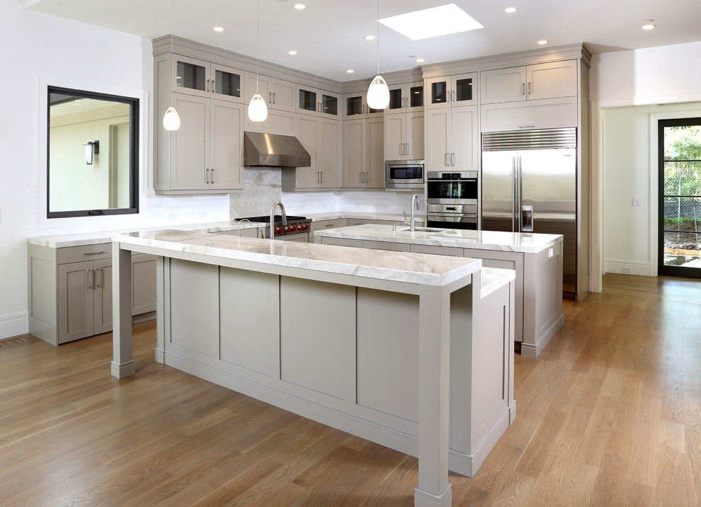 Modern inredning av ett stort kök, med en undermonterad diskho, skåp i shakerstil, grå skåp, marmorbänkskiva, vitt stänkskydd, stänkskydd i stenkakel, rostfria vitvaror, ljust trägolv och flera köksöar