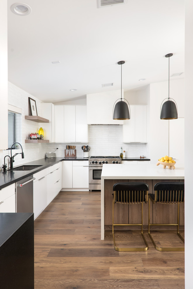 Inspiration for a contemporary l-shaped kitchen in Phoenix with a submerged sink, flat-panel cabinets, white cabinets, white splashback, stainless steel appliances, medium hardwood flooring, an island, brown floors and black worktops.