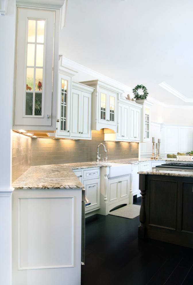 Klassisk inredning av ett stort kök, med en rustik diskho, luckor med profilerade fronter, vita skåp, granitbänkskiva, grått stänkskydd, mörkt trägolv och en köksö