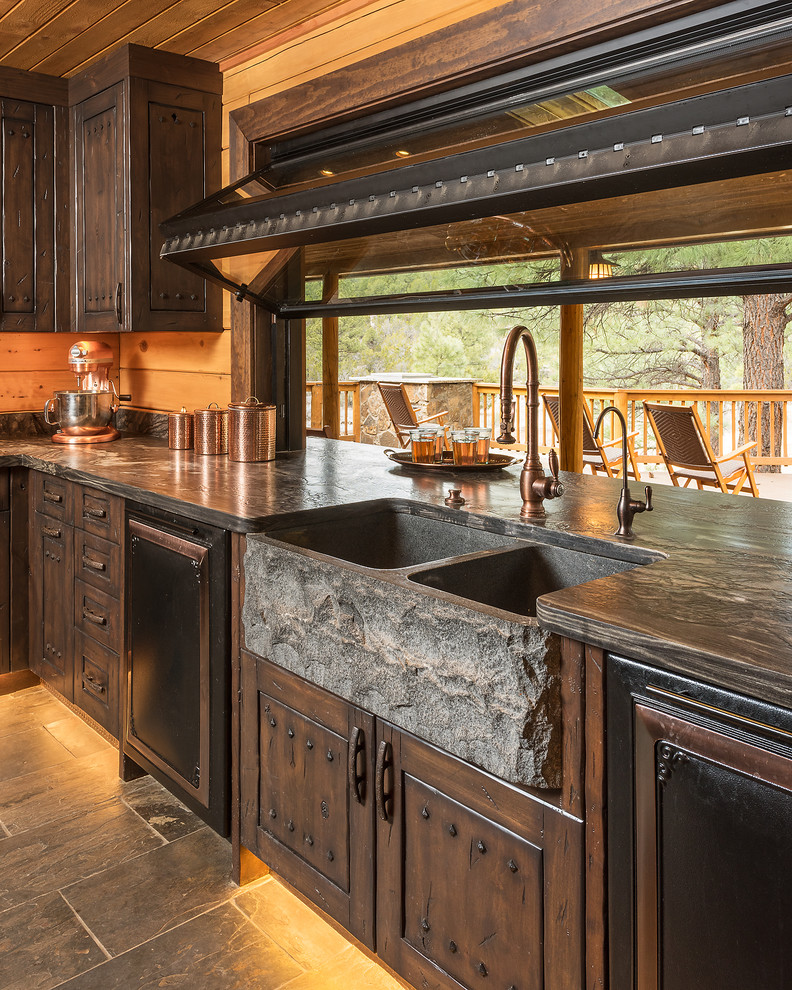 Inredning av ett rustikt stort kök med öppen planlösning, med en rustik diskho, luckor med upphöjd panel, bruna skåp, bänkskiva i kalksten, beige stänkskydd, stänkskydd i trä, svarta vitvaror, skiffergolv och en köksö