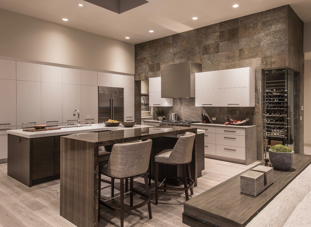 This is an example of an expansive contemporary u-shaped open plan kitchen in Phoenix with a submerged sink, flat-panel cabinets, white cabinets, engineered stone countertops, slate splashback, stainless steel appliances, light hardwood flooring, multiple islands, beige floors and grey worktops.