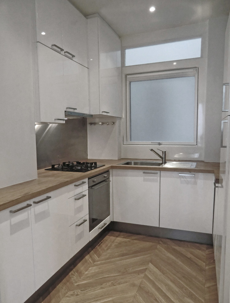 Cette image montre une cuisine minimaliste en U avec un évier 1 bac, un placard à porte plane, des portes de placard blanches, un plan de travail en bois, une crédence métallisée et un électroménager noir.