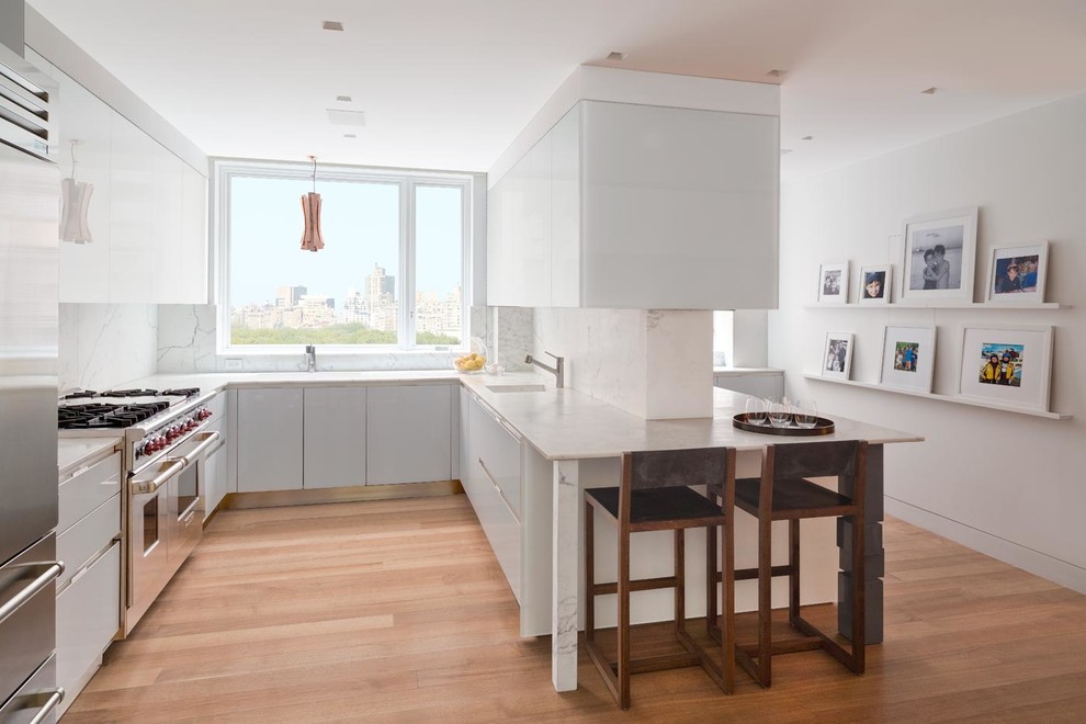Foto på ett funkis u-kök, med släta luckor, vita skåp, vitt stänkskydd, stänkskydd i sten, rostfria vitvaror, mellanmörkt trägolv och en halv köksö