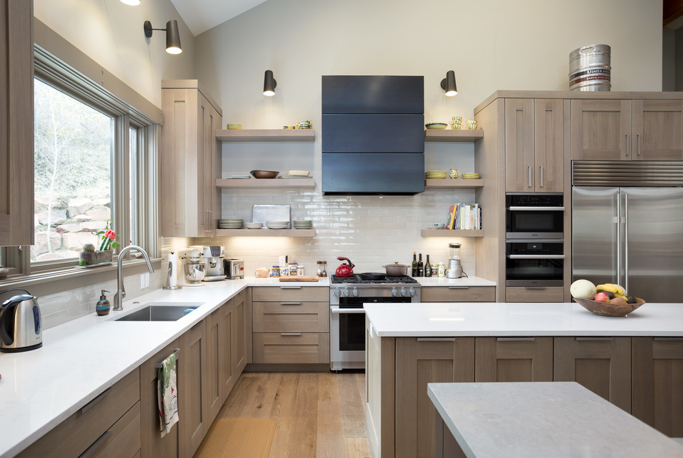 This is an example of a large contemporary galley open plan kitchen in Salt Lake City with a submerged sink, shaker cabinets, beige cabinets, wood worktops, white splashback, ceramic splashback, stainless steel appliances, light hardwood flooring, multiple islands, beige floors and white worktops.
