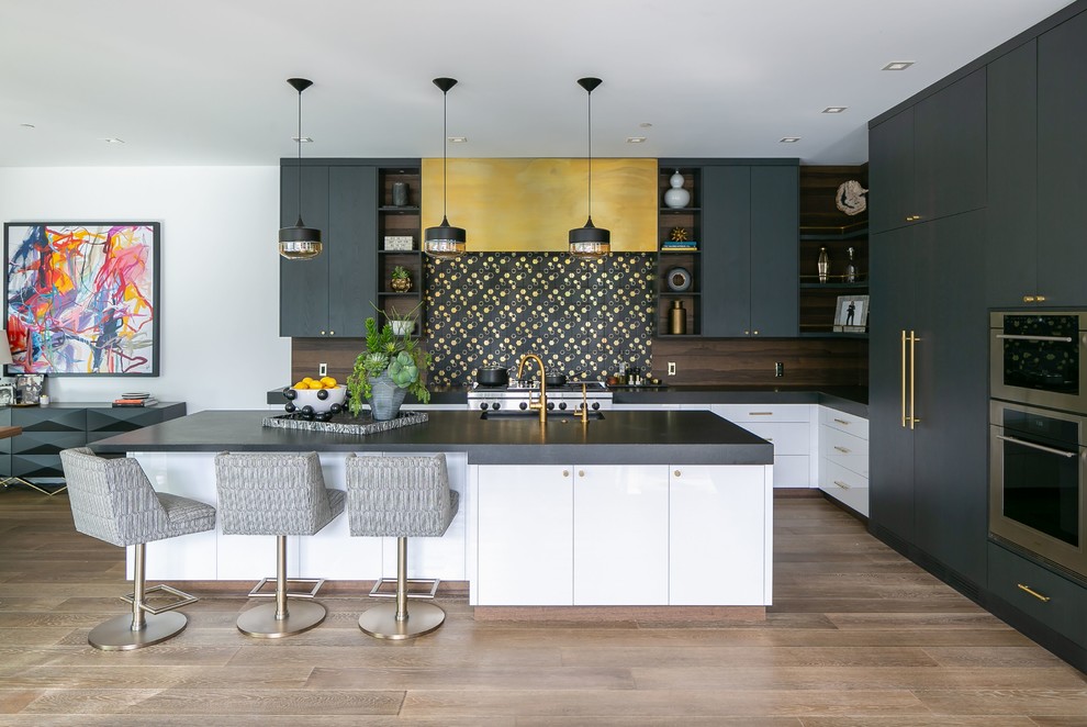 Modern inredning av ett stort svart svart l-kök, med en undermonterad diskho, släta luckor, svarta skåp, flerfärgad stänkskydd, rostfria vitvaror, mellanmörkt trägolv, en köksö och brunt golv
