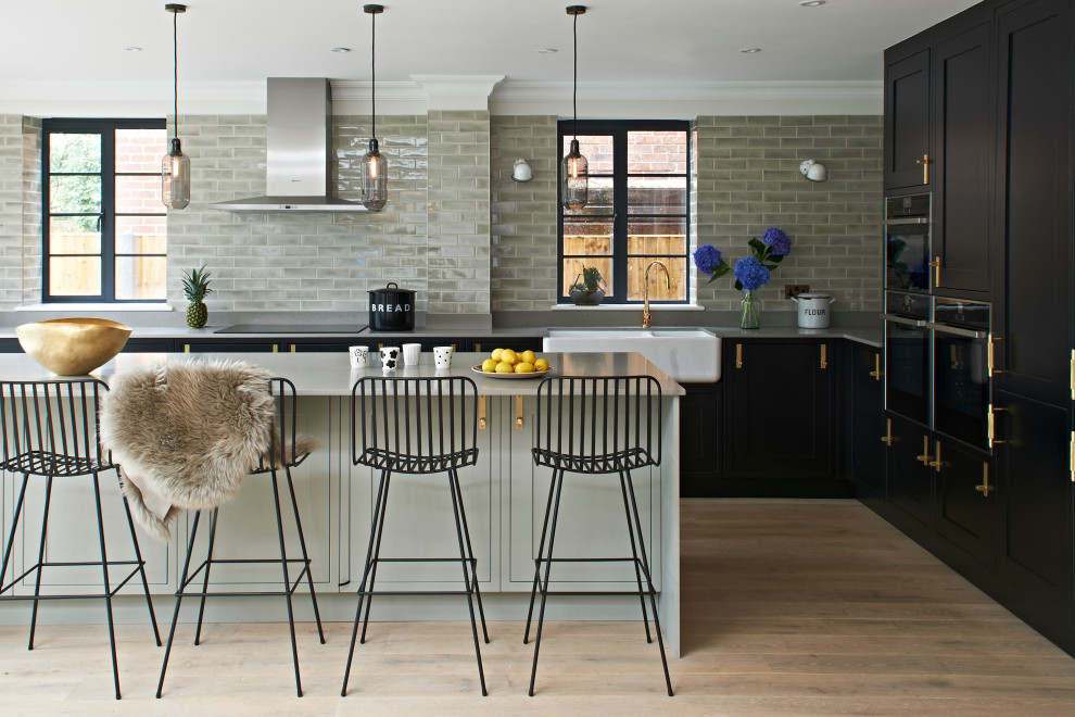 Photo of a traditional u-shaped open plan kitchen in Hertfordshire with a belfast sink, shaker cabinets, black cabinets, grey splashback, metro tiled splashback, black appliances, medium hardwood flooring, an island, brown floors and grey worktops.