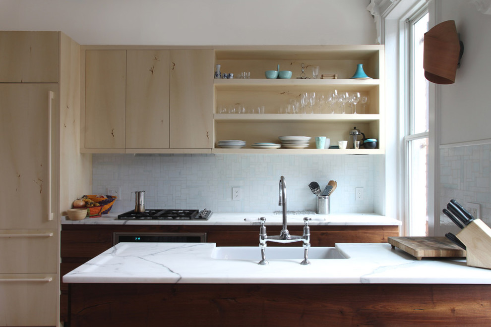 Modelo de cocina contemporánea con encimera de mármol, fregadero sobremueble, armarios abiertos, puertas de armario de madera clara, salpicadero blanco y electrodomésticos con paneles