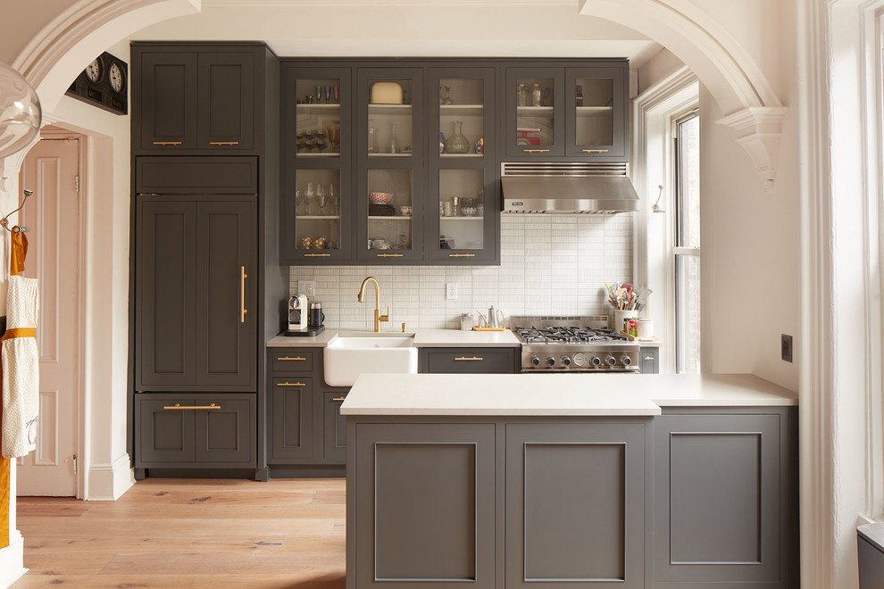 This is an example of a large classic single-wall open plan kitchen in New York with a belfast sink, grey cabinets, white splashback, stainless steel appliances, a breakfast bar, engineered stone countertops, matchstick tiled splashback, light hardwood flooring, beige floors and recessed-panel cabinets.