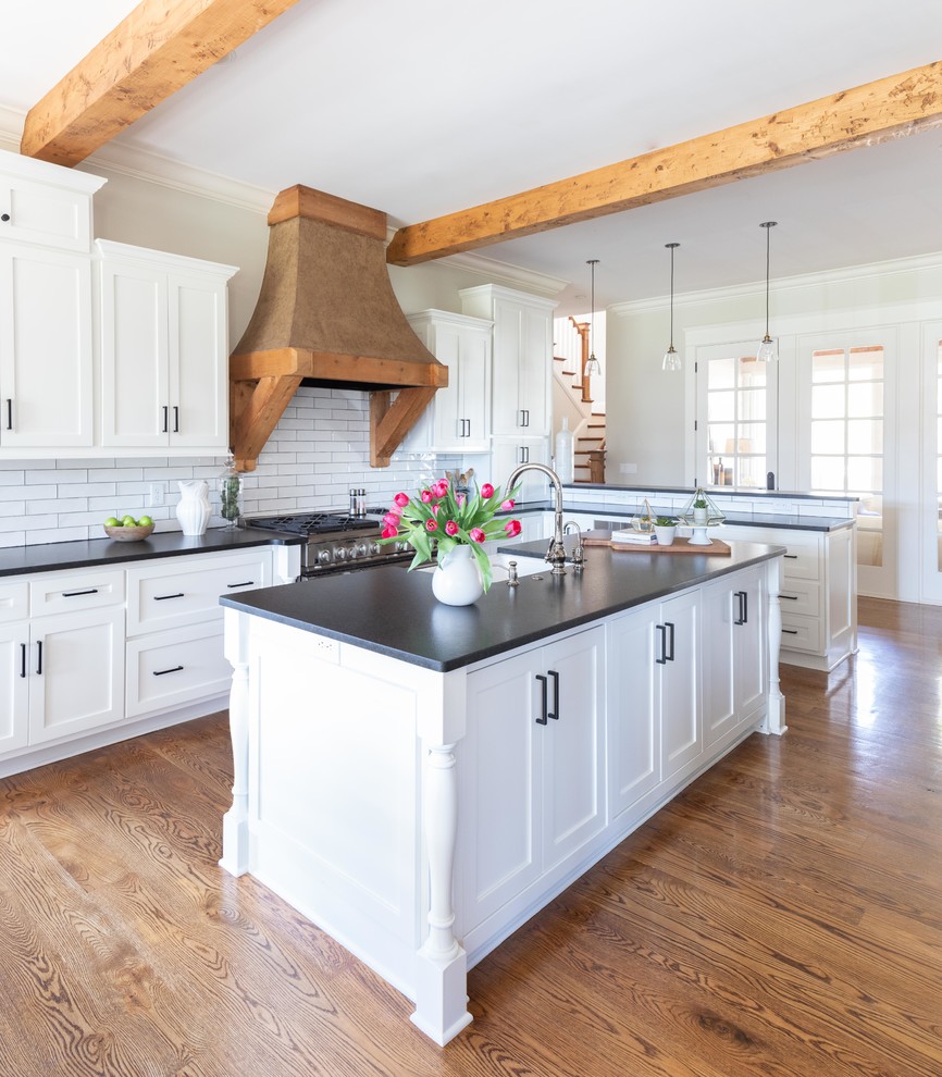 This is an example of a large classic l-shaped kitchen/diner in Nashville with a belfast sink, shaker cabinets, white cabinets, white splashback, metro tiled splashback, stainless steel appliances, medium hardwood flooring, an island, brown floors, black worktops and engineered stone countertops.