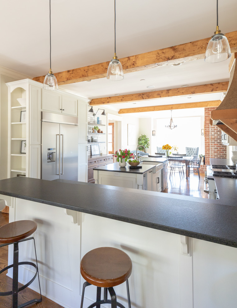 Imagen de cocina comedor tradicional renovada de tamaño medio con fregadero sobremueble, armarios estilo shaker, puertas de armario blancas, encimera de cuarzo compacto, salpicadero blanco, salpicadero de azulejos de cerámica, electrodomésticos de acero inoxidable, suelo de madera en tonos medios, una isla, suelo marrón y encimeras negras