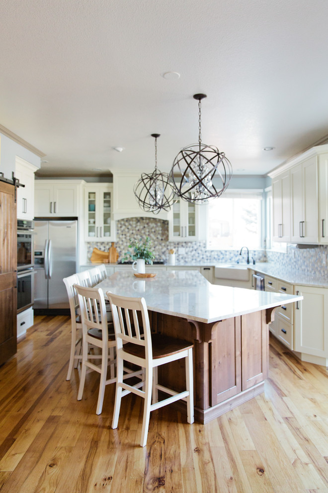 Photo of a large traditional u-shaped kitchen/diner in Denver with shaker cabinets, white cabinets, engineered stone countertops, multi-coloured splashback, an island, a belfast sink, porcelain splashback, stainless steel appliances and light hardwood flooring.