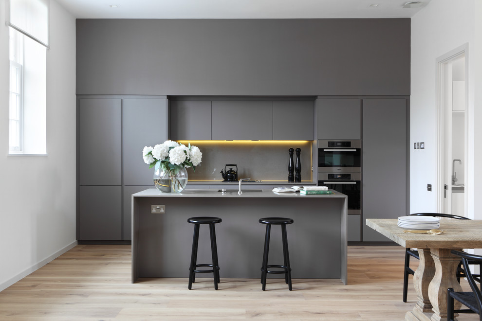 Modern inredning av ett kök och matrum, med en enkel diskho, släta luckor, grå skåp, grått stänkskydd, rostfria vitvaror, ljust trägolv och en köksö