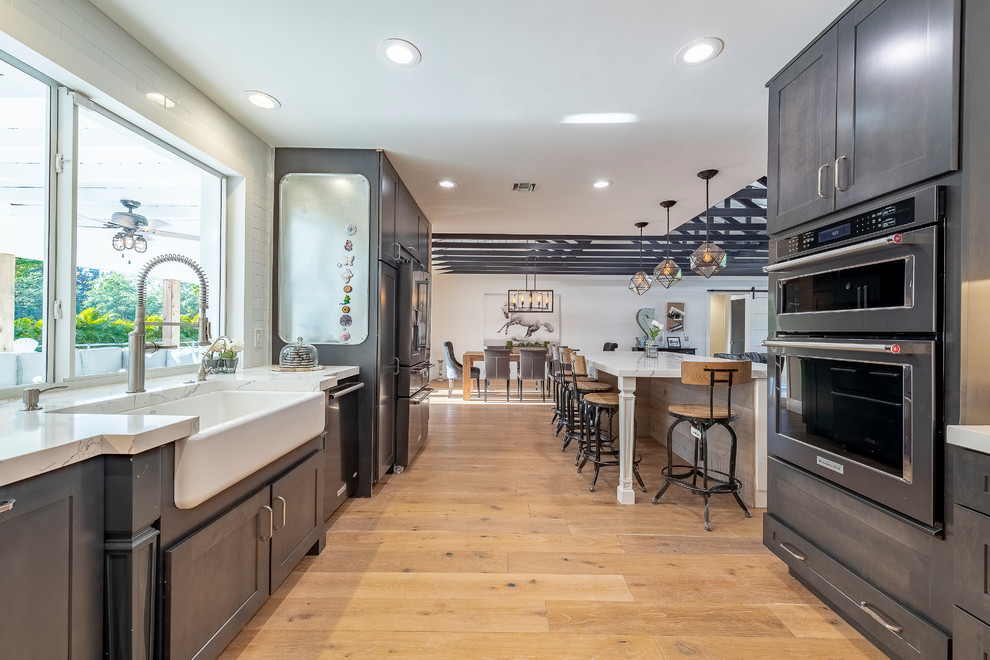 Photo of a rural kitchen in Miami with a belfast sink, shaker cabinets, white cabinets, composite countertops, black appliances, light hardwood flooring and an island.