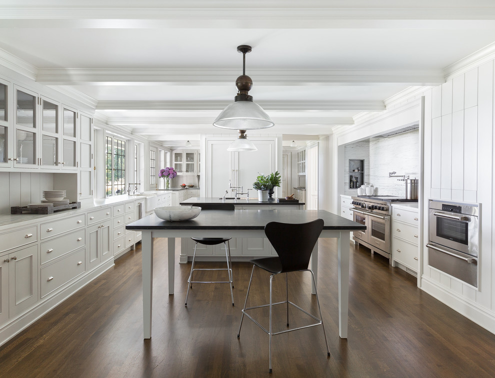 Traditional kitchen in Seattle with a belfast sink, glass-front cabinets, white cabinets, white splashback, stainless steel appliances, dark hardwood flooring, multiple islands and brown floors.