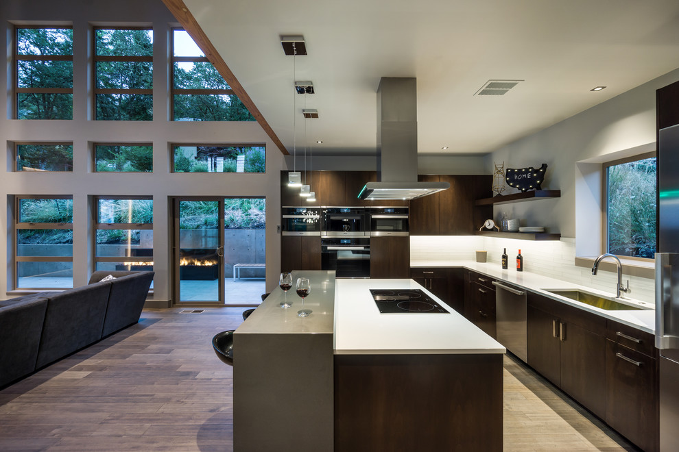 Cette image montre une cuisine ouverte design en bois foncé avec un évier encastré, un placard à porte plane, une crédence blanche, un électroménager en acier inoxydable, un sol en bois brun et îlot.
