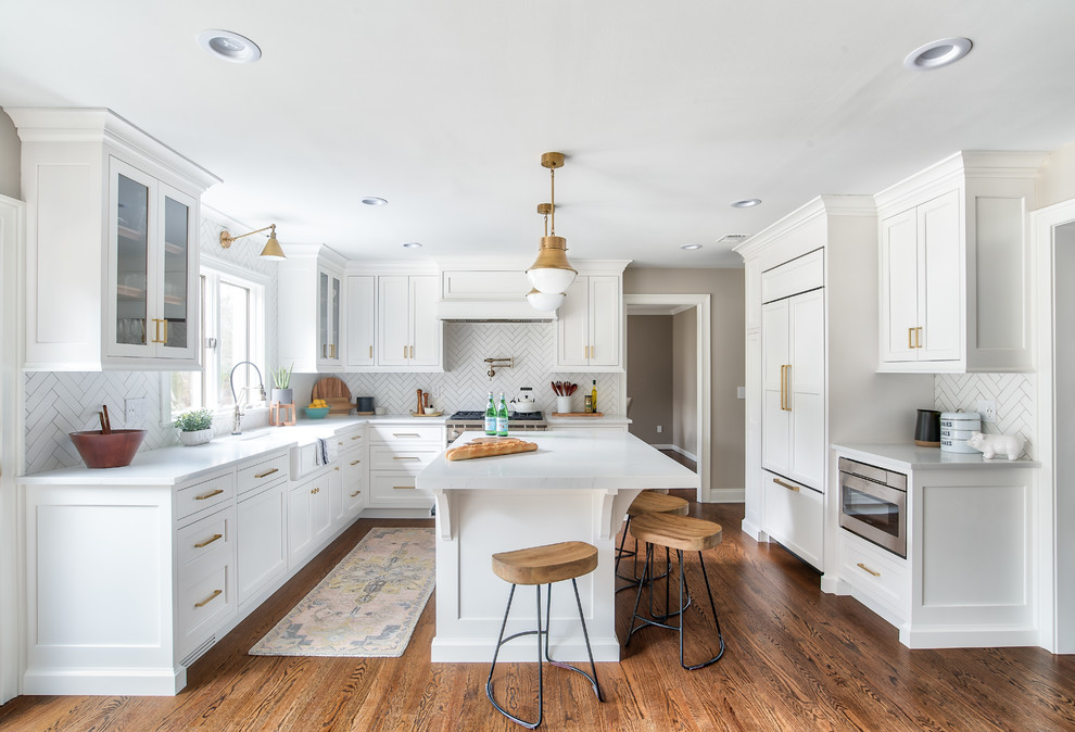 Traditional u-shaped kitchen in New York with a belfast sink, white cabinets, engineered stone countertops, integrated appliances, shaker cabinets, white splashback, medium hardwood flooring, an island, brown floors and white worktops.